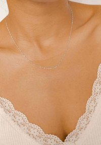 Collana in argento con un delicato design a catena, poggiata sulla clavicola, abbinata a un indumento con rifiniture in pizzo bianco.