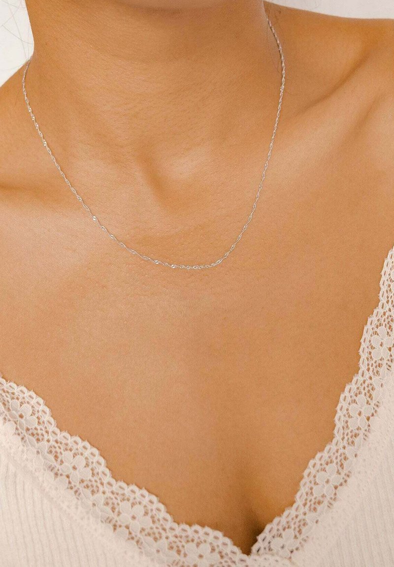 Collana in argento con un delicato design a catena, poggiata sulla clavicola, abbinata a un indumento con rifiniture in pizzo bianco.