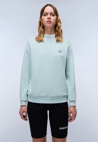 Felpa verde menta con collo a giro, polsini e orlo a costine, abbinata a pantaloni bicycle neri con dettaglio del logo sul lato.