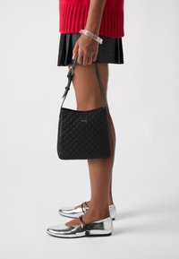 Borsa a tracolla nera trapuntata, con superficie liscia e logo. Indossata con un maglione rosso e ballerine argento.