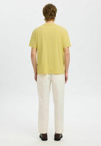 T-shirt en coton jaune à manches courtes, col rond, associé à un pantalon droite de couleur crème et des chaussures marron foncé, vu de derrière.