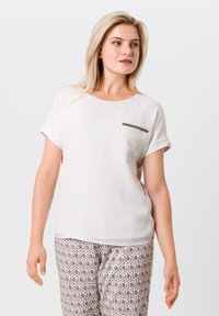 Helles beiges Kurzarmblusenshirt mit Rundhalsausschnitt und einer Tasche mit gestreiftem Detail. Kombiniert mit gemusterten beigen Hosen.