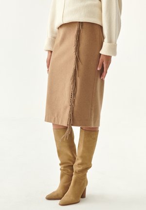 Woman wearing a tan knee-length skirt with fringe detail and tan suede heeled boots, paired with a cream sweater.