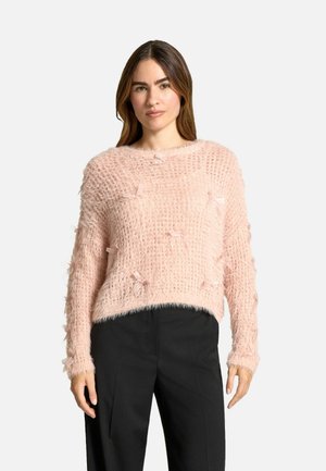 Maglione lungo a maniche lunghe di colore rosa, realizzato in materiale soffice e con una texture, caratterizzato da piccoli fiocchi e una vestibilità ampia, abbinato a pantaloni neri.