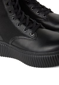 Czarne wysokie trampki z teksturowaną tkaniną, płaskimi sznurowadłami i grubą, żłobioną podeszwą. Posiada metalowe oczka i wytłoczone logo.