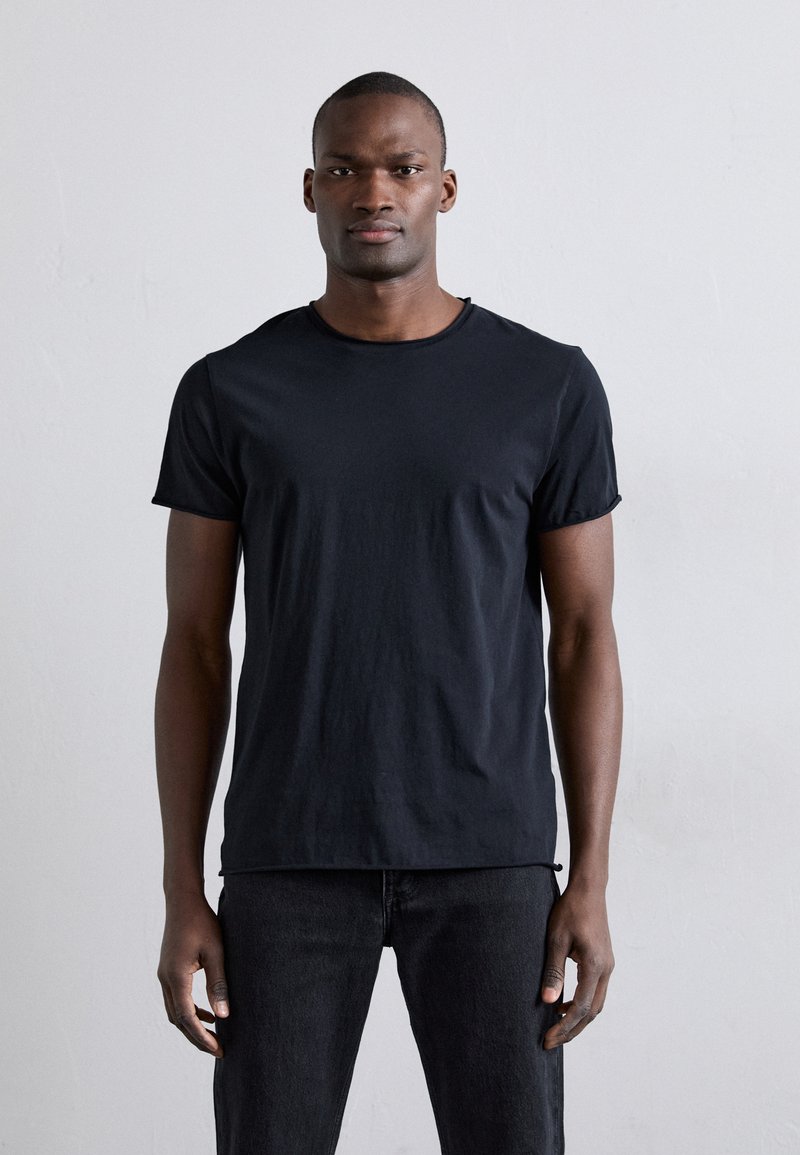 Camiseta negra de manga corta con cuello redondo. Hecha de una tela suave, tiene una forma ajustada y una textura lisa. Se lleva con jeans oscuros.