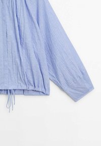 Blouse bleu clair texturée à manches longues, ourlet élastique et cordons décoratifs à la taille. Tissu léger avec une coupe décontractée.