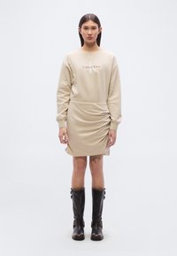 Μπεζ φόρεμα-φ ούτερ Calvin Klein με μακριά μανίκια, σχεδιασμό με ρυτίδες στο πλάι και ορατό λογότυπο στο στήθος. Συνδυάζεται με καφέ μπότες μέχρι το γόνατο.