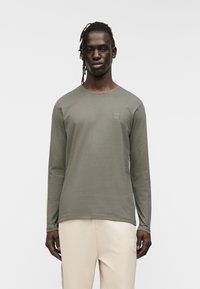 Πουκάμισο ελιάς με μακριά μανίκια από ελαφρύ ύφασμα, με στρογγυλή λαιμόκοψη και μικρό λογότυπο στο στήθος. Απλός, εφαρμοστός σχεδιασμός.