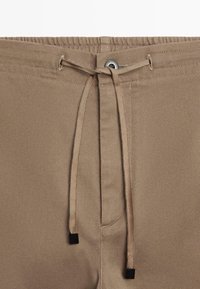 Brązowy, teksturowany pas z elastycznym ściągaczem i sznurkami; wyposażony w metalowy guzik i przeszycia dekoracyjne. Zaprojektowany dla luźnego kroju.