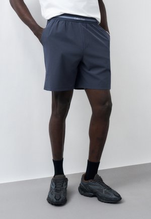 Homme portant un short bleu marine, des chaussettes noires et des chaussures de sport noires, debout contre un mur gris clair, les mains dans les poches.