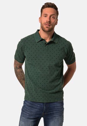 Polo vert avec un motif texturé de petits diamants noirs. Il présente un col et des manches courtes, associé à un jean en denim bleu.