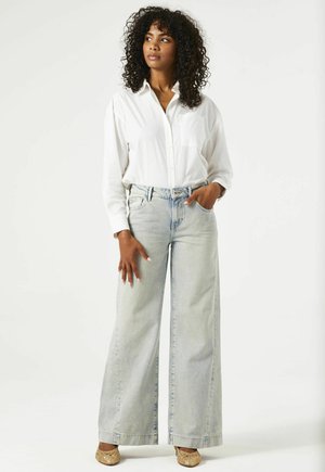 Chemise blanche à boutons, jean en denim bleu clair, large et détendu, avec un détail de poche, et accentué par des chaussures tressées couleur tan.