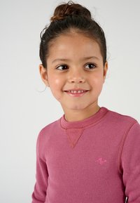 Camicia a maniche lunghe in rosa morbido, con texture a coste, dotata di scollatura rotonda e cuciture a contrasto, con un piccolo logo sul petto.