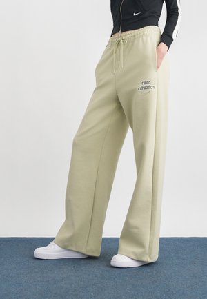 Άτομο που φοράει μπεζ φαρδιά παντελόνια φόρμας με το λογότυπο "nike athletics", μαύρο μπουφάν με φερμουάρ, λευκά αθλητικά παπούτσια, στέκεται σε μπλε χαλί.