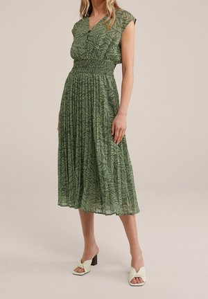 Grünes Midi-Kleid mit V-Ausschnitt, gerafftem Taillenbund, kurzärmeligen Cap-Ärmeln und einem Blättermuster. Kombiniert mit hellgrünen Slip-On-Heels.