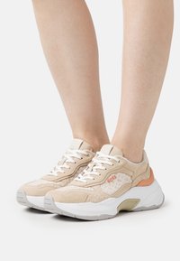 Beiga mocka- och mesh-sneakers med vita snören, orange BOSS-logga och tjock vit sula, barfota.