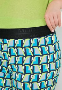 Primer plano de la mano de una persona apoyada en la cadera, vistiendo pantalones con un patrón geométrico en azul, verde azulado y amarillo, y una cintura negra.