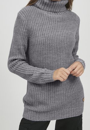 Frau trägt einen grauen grob gestrickten Rollkragenpullover mit langen Ärmeln, hält die Hände vor sich zusammen, vor einem einfarbigen Hintergrund.