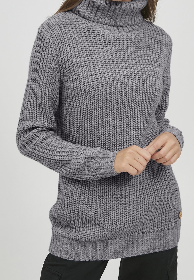 Femme portant un pull à col roulé en maille épaisse grise à manches longues, tenant ses mains jointes devant elle, sur un fond uni.