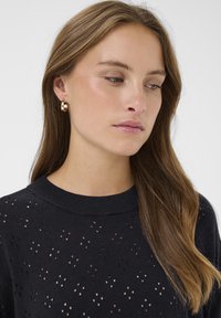 Pull en tricot bleu marine avec un design à trous ouverts, doté d'un col rond. De grandes boucles d'oreilles en or avec une accent perle sont visibles.