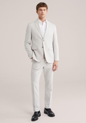 Mladý muž stojaci, oblečený v svetlobéžovom obleku, bielej košeli a čiernych spoločenských topánkach, na neutrálnom jednoduchom pozadí.