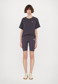 Sötét szürke, rövidujjú póló, kicsi logóval, összeillő kerékpáros sorttal párosítva. A modellen világos sportcipők vannak, texturált talppal.