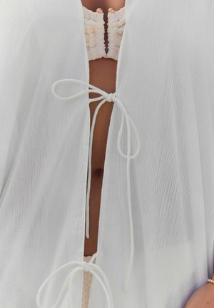 Copertura leggera bianca con tessuto testurizzato, scollatura a V e chiusura con cordoncino sul davanti. Accentuata da un top interno arricciato.
