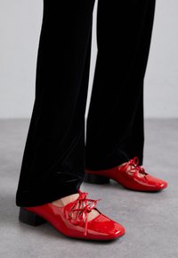 Red patent leather shoes with a square toe, featuring a bow tie detail and low block heel. Paired with black velvet pants.