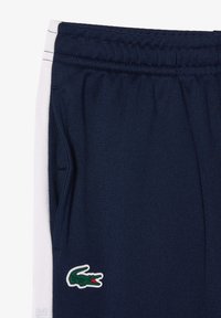 Granatowe szorty sportowe z białymi paskami po bokach, elastyczną talią, boczną kieszenią i zielonym logo krokodyla blisko dołu.