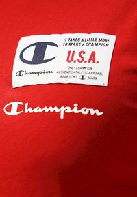 T-shirt in cotone rosso con logo bianco "Champion" e un'etichetta con testo blu e rosso, che indica "U.S.A." e i dettagli del marchio.