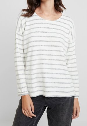 Kvinde iført en hvid langærmet skjorte med tynde grå vandrette striber og mørkegrå jeans mod en ensfarvet baggrund.