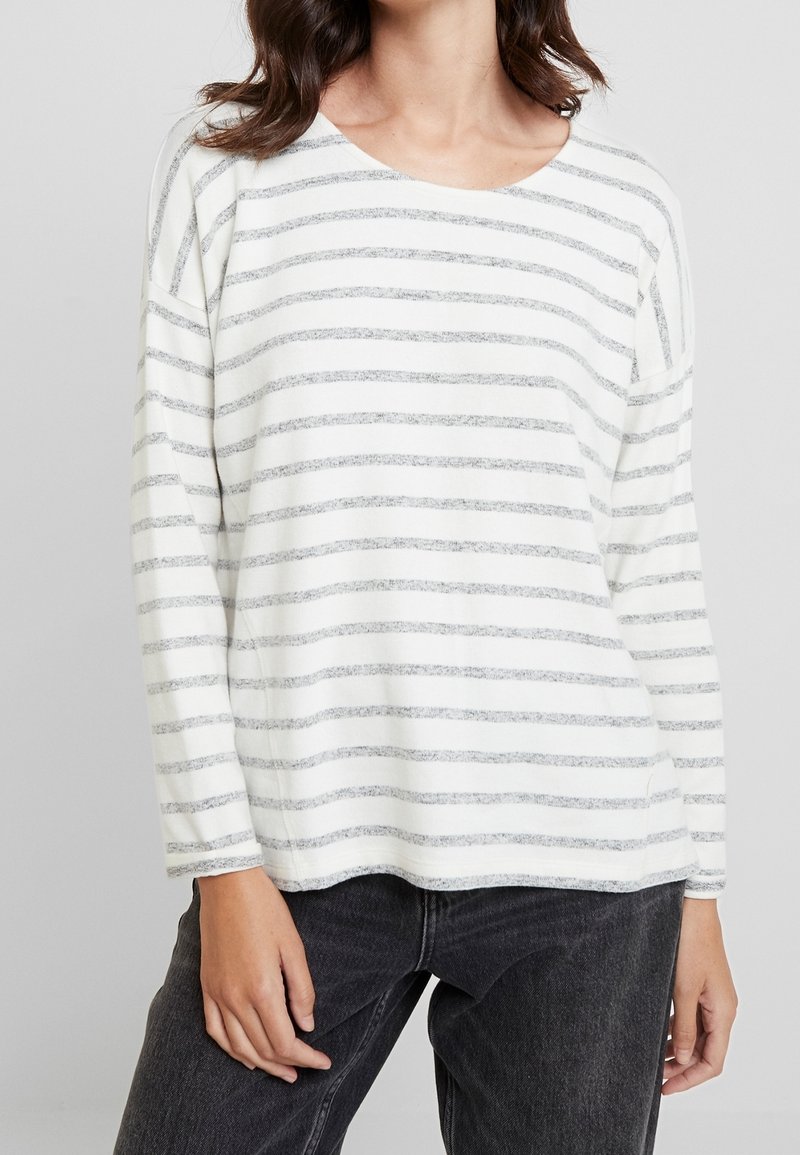 Femme portant une chemise blanche à manches longues avec de fines rayures horizontales grises et un jean gris foncé sur un fond uni.