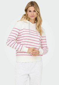 Donna con capelli biondi che indossa un hoodie bianco con strisce orizzontali rosa e pantaloni della tuta grigi chiari, su uno sfondo neutro.