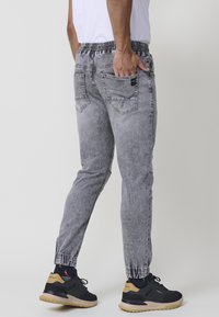 Pantalones jogger de mezclilla gris con cintura elástica, bolsillos laterales, piernas estrechas y un acabado desgastado. Combinados con zapatillas negras.