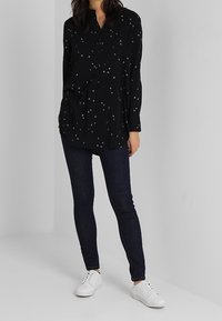 Chemise noire boutonnée avec des motifs en forme d'étoiles, associée à un jean skinny foncé et des baskets blanches, créant un contraste de couleur et de texture.