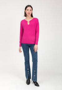 Heldroze sweater met een keyhole-halslijn, gecombineerd met blauwe denim flared jeans met knoopaccenten aan de zoom en zwarte hakken.