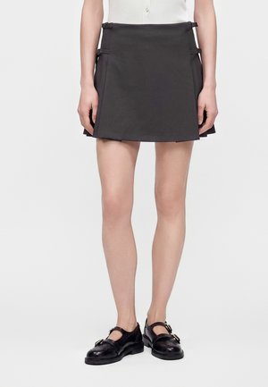 Femme portant une jupe plissée courte noire avec des détails à boucle et des chaussures Mary Jane noires, debout sur un fond blanc.