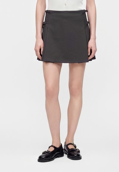 Femme portant une jupe plissée courte noire avec des détails à boucle et des chaussures Mary Jane noires, debout sur un fond blanc.