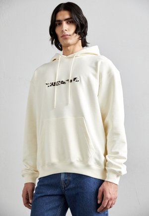 Cream-colored hoodie with a front kangaroo pocket, drawstrings, and a textured graphic logo in dark tones across the chest. Paired with blue jeans.