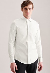 Camicia bianca a maniche lunghe con colletto strutturato, chiusura frontale con bottoni e silhouette aderente. Tessuto liscio, abbinata a pantaloni neri.