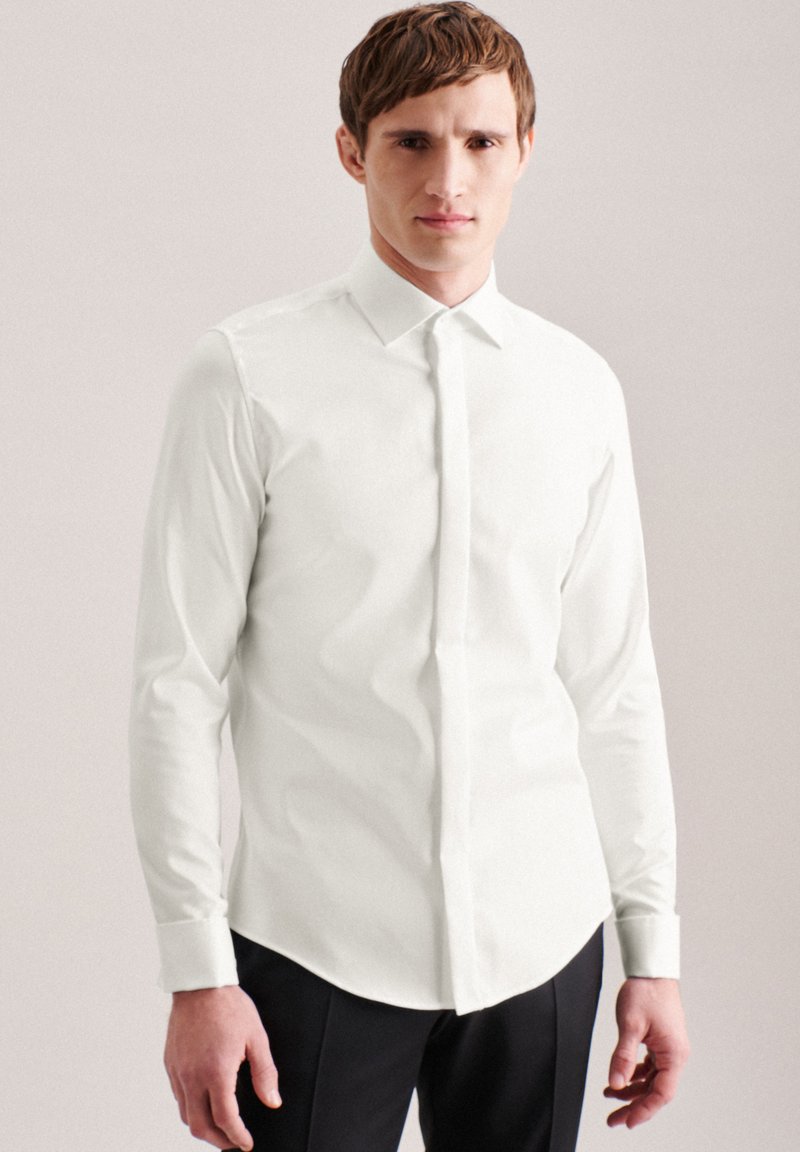 Camicia bianca a maniche lunghe con colletto strutturato, chiusura frontale con bottoni e silhouette aderente. Tessuto liscio, abbinata a pantaloni neri.