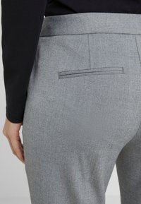 Close-up van een persoon die lichtgrijze broek draagt met een achterzak en een zwart shirt met lange mouwen, hand rustend naast de heup.
