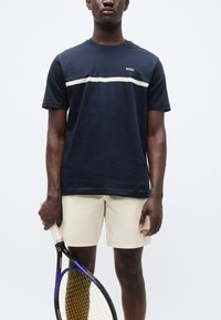 Μπλε ναυτικό βαμβακερό μπλουζάκι με οριζόντια λευκή ρίγα και λογότυπο "BOSS", συνδυασμένο με μπεζ σορτς που φέρουν κεντημένο λογότυπο. Κρατά ρακέτα του τένις.