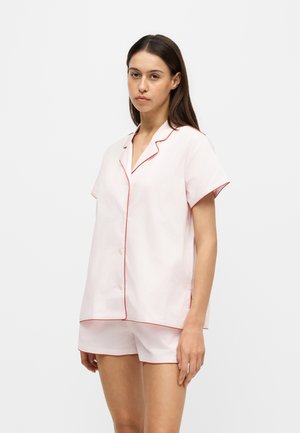 Femme aux longs cheveux foncés portant une chemise légère rose à manches courtes boutonnée et un short assorti avec une bordure rouge sur fond blanc.