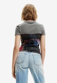 Grå kortærmet bomuldsbluse med en farverig, mønstret rynket ryg og sort bånd, parret med lyseblå posede jeans.