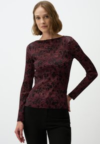 Langarmshirt in kastanjebruin met een gestructureerd zwart patroon, voorzien van een bootnek en een fitted silhouet. Draagt met zwarte broek.