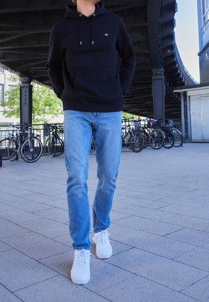 Person trägt schwarzen Hoodie, hellblaue Jeans und weiße Turnschuhe und läuft auf einem gefliesten Bürgersteig in der Nähe von Fahrrädern unter einer Metallbrücke.