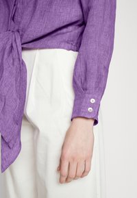 Gros plan sur une personne portant une chemise à manches longues violette avec des poignets boutonnés et un nœud à l'avant, associée à un pantalon blanc taille haute.