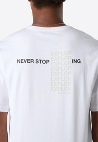 Mand iført hvid T-shirt med teksten "NEVER STOP EXPLORING" i sort og falmet tryk på ryggen, stående mod en ensfarvet baggrund.
